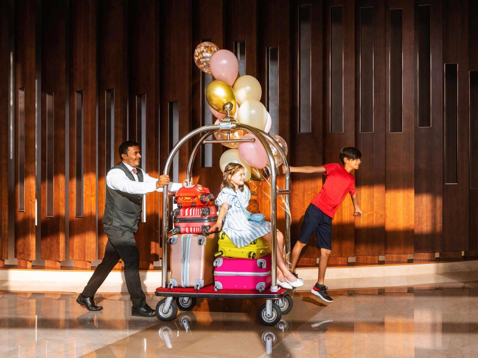 สวิสโซเทล อัล กูร์เร Hotel ดูไบ ภายนอก รูปภาพ The photo depicts a hotel scene where a bellhop is pushing a luggage cart. The cart is piled high with various colorful suitcases, and it is topped with balloons in shades of pink, gold, and white. On the cart sits a girl dressed in a light blue dres